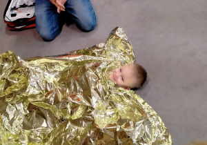 Chłopiec okryty kocem termicznym, leży na dywanie.