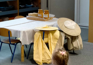 Na stole stoi taca z waflami i miodami. Na krzesełkach wiszą stroje pszczelarskie.